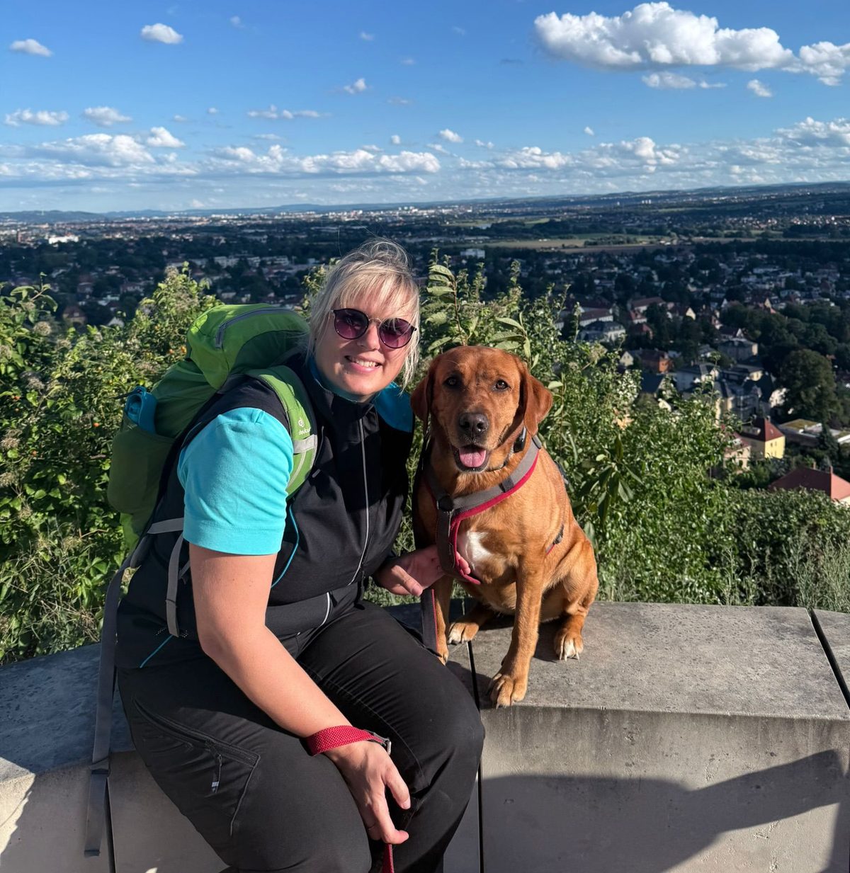 Antje mit ihrem Hund auf einer Mauer an einem Aussichtspunkt, im Hintergrund eine weite Sicht über Stadt und Umland